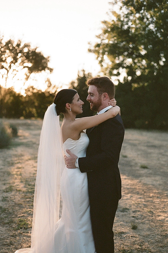 Couple portrait of bride and groom embrace at golden-hour outdoors, her long veil and strapless dress flowing beside his black tuxedo in a field at sunset