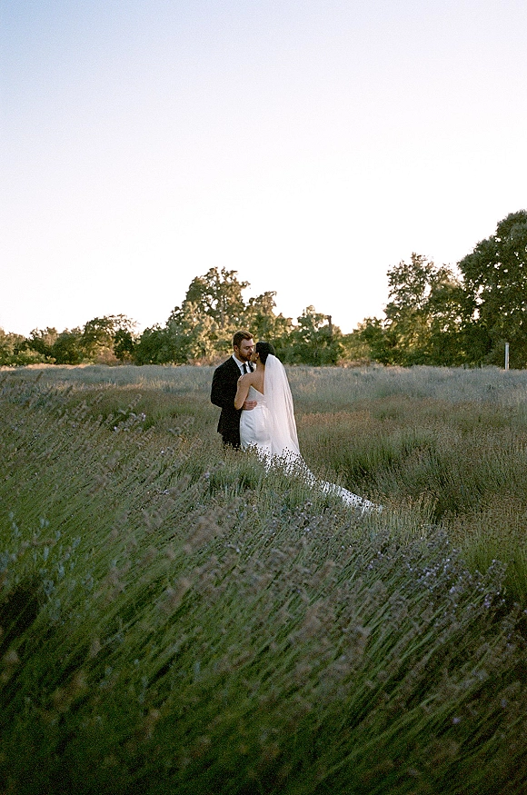 Couple portrait in a wildflower field, bride and groom embrace in tall grass with a flowing bridal veil under open sky