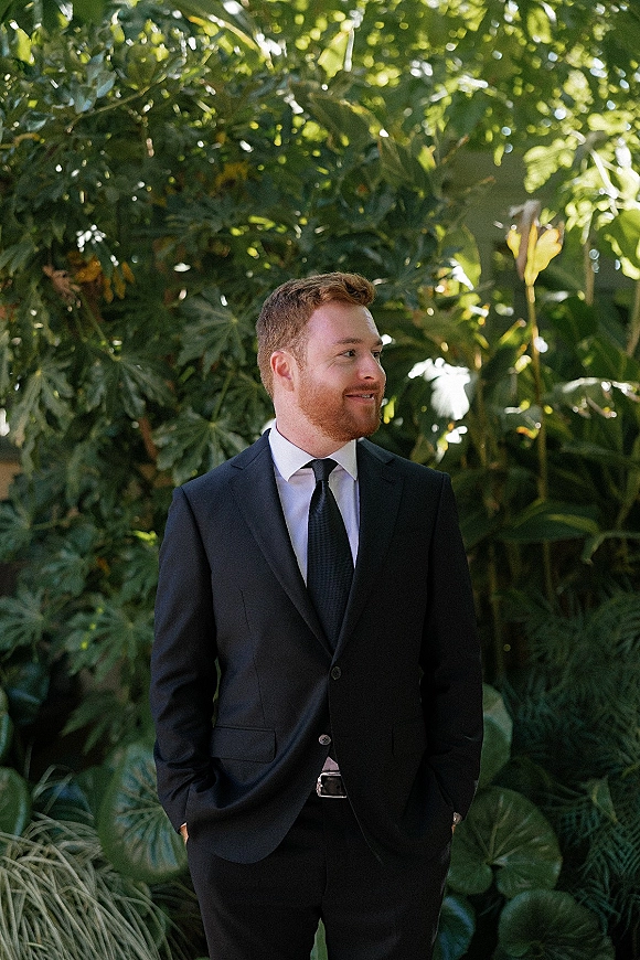 Groom portrait of a redhead groom in black suit with hands in pockets, looking away against lush tropical garden greenery in dappled sun