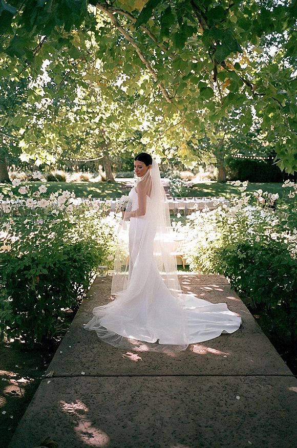 Bridal portrait of a bride in a long veil holding a white bouquet, looking over her shoulder on a garden stone walkway under string lights