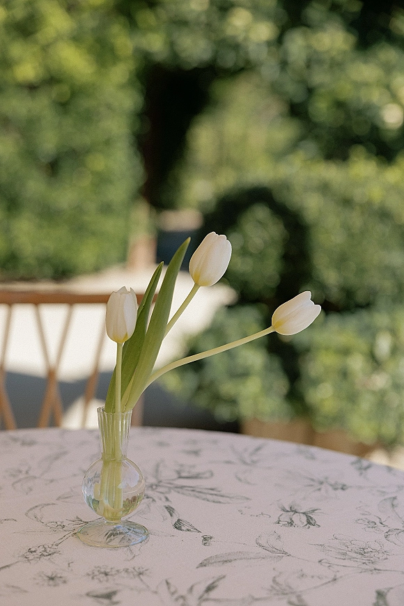 Wedding table centerpiece with a tulip bud vase holding a single white stem on a round printed tablecloth, set on an outdoor patio with greenery