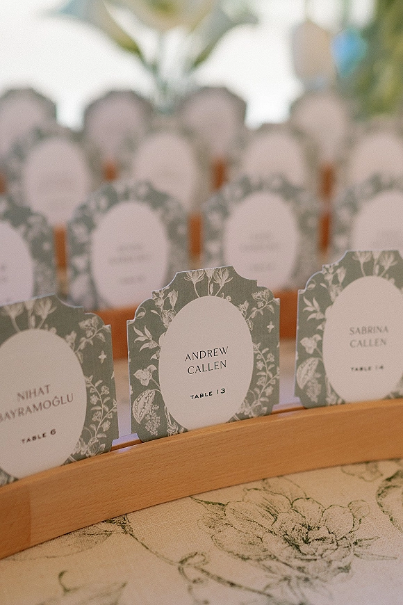 Wedding place cards and wedding escort cards with botanical illustrations in a wooden holder on a table, with a softly blurred display behind