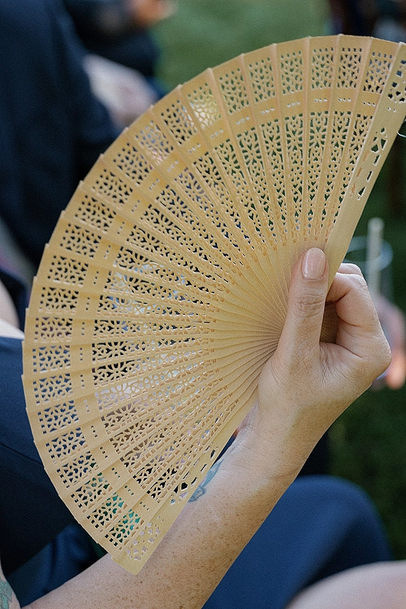 Wedding ceremony fan, a folding hand fan with lace cutout pattern held by a guest with nude manicure amid outdoor greenery seating