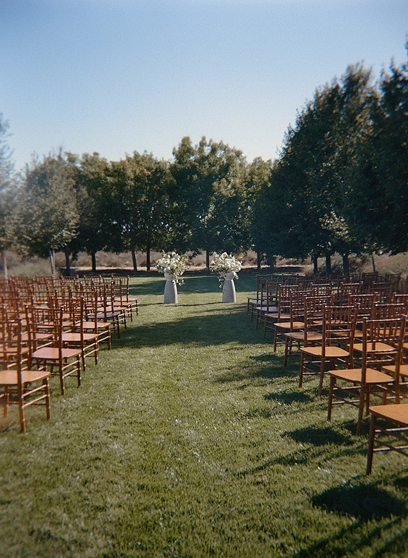 Ceremony setup for an outdoor wedding ceremony with wood chiavari chairs and white floral pedestal vases on a green lawn under blue sky