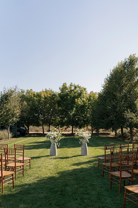 Ceremony setup for an outdoor wedding ceremony with rows of wood Chiavari chairs and floral pedestal vases on a grass lawn under trees