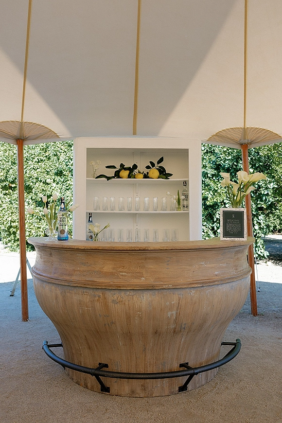 Wedding bar setup with a rustic wooden bar, glassware and liquor bottles, plus lemon and calla lily decor under a white tent canopy