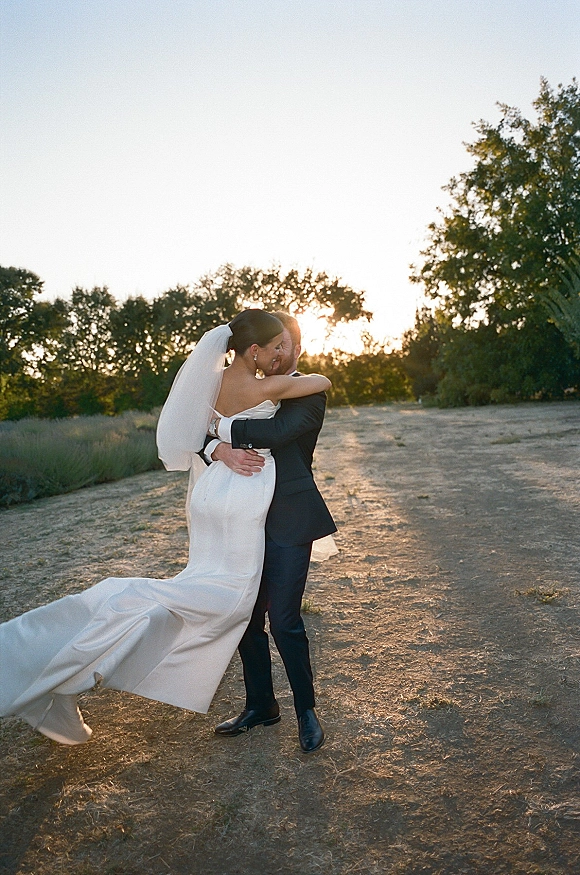 Wedding kiss portrait of bride and groom kissing on a dirt path in a field at sunset, her veil and long dress train flowing