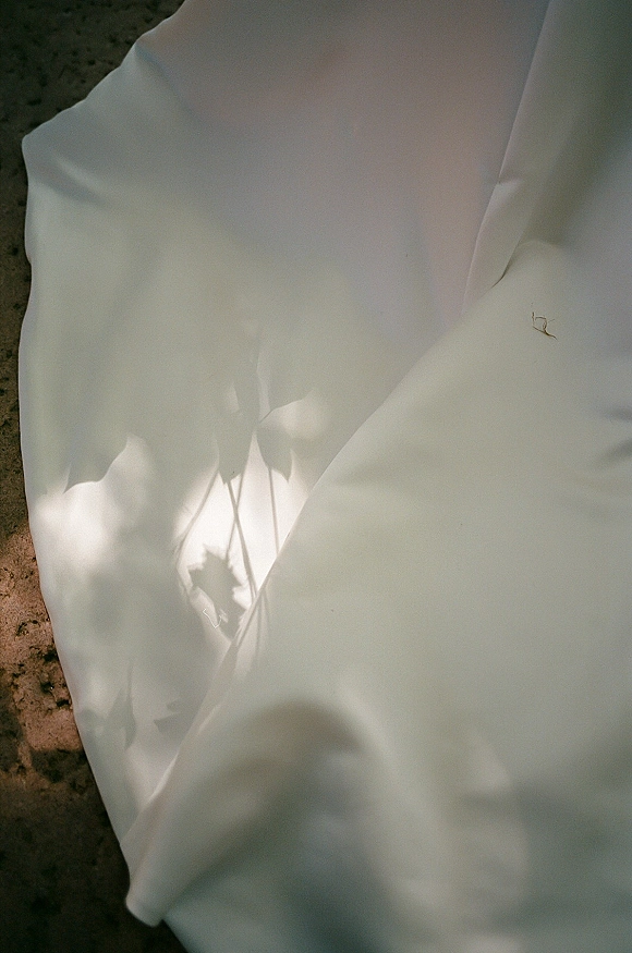 Wedding dress close-up showing bridal gown fabric detail in satin folds, hem and train draping on sunlit stone ground with leaf shadows