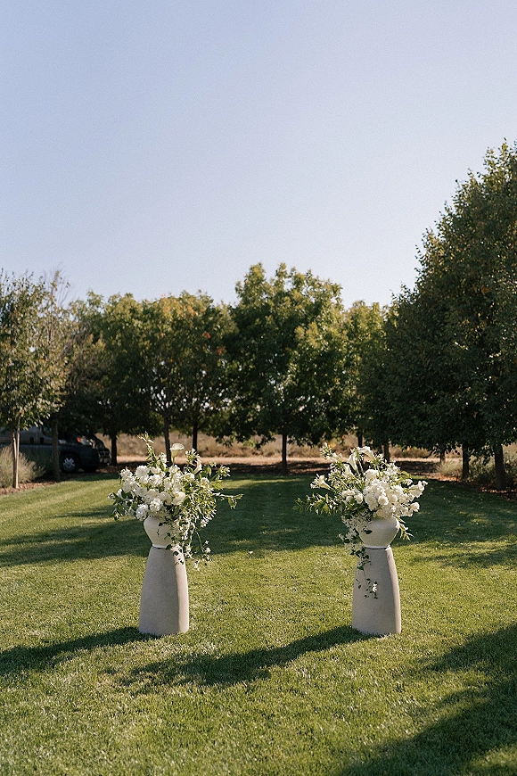 Ceremony aisle decor with white floral arrangements and greenery on pedestal stands, lining a grass lawn under trees and blue sky