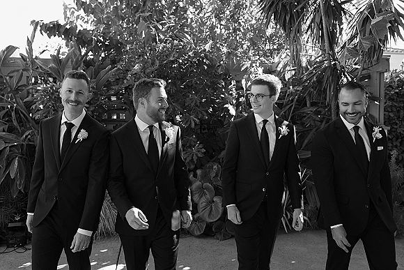 Groomsmen portrait of men walking together in black suits with boutonnières, laughing along a sunlit outdoor walkway with tropical foliage behind