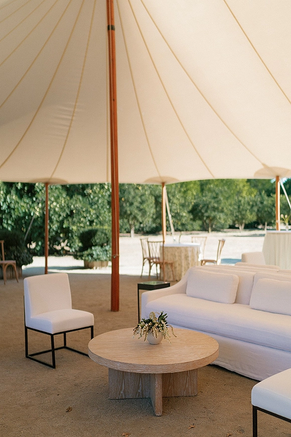 Wedding lounge setup with a white sofa and upholstered chair around a round wood coffee table and floral centerpiece under a tent canopy