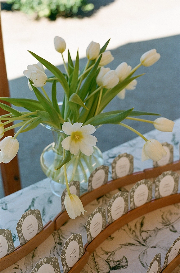 Wedding escort cards in wooden holders beside a clear glass vase of white tulips on a patterned tablecloth on an outdoor patio