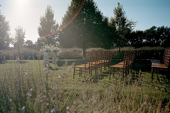 Outdoor ceremony setup with wood chairs and white floral pedestal arrangements lining the aisle on a sunny lawn beside a wildflower field