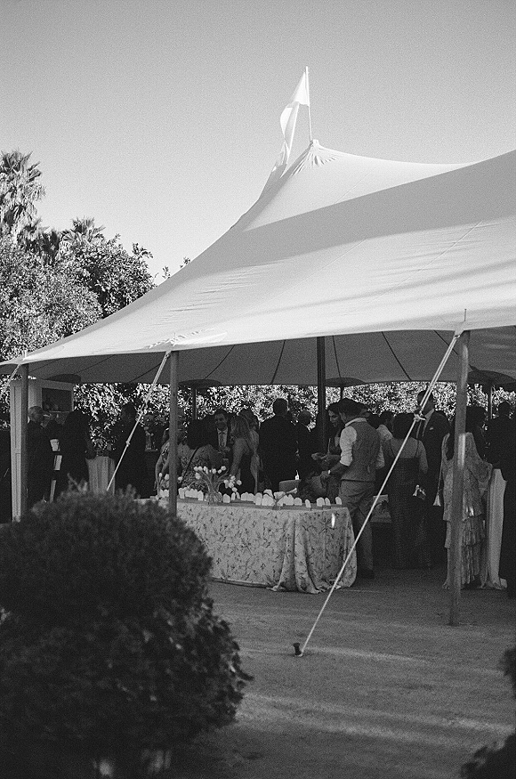 Wedding cocktail hour under an outdoor wedding tent with guests mingling around linen cocktail tables, glassware, and floral vases on a lawn