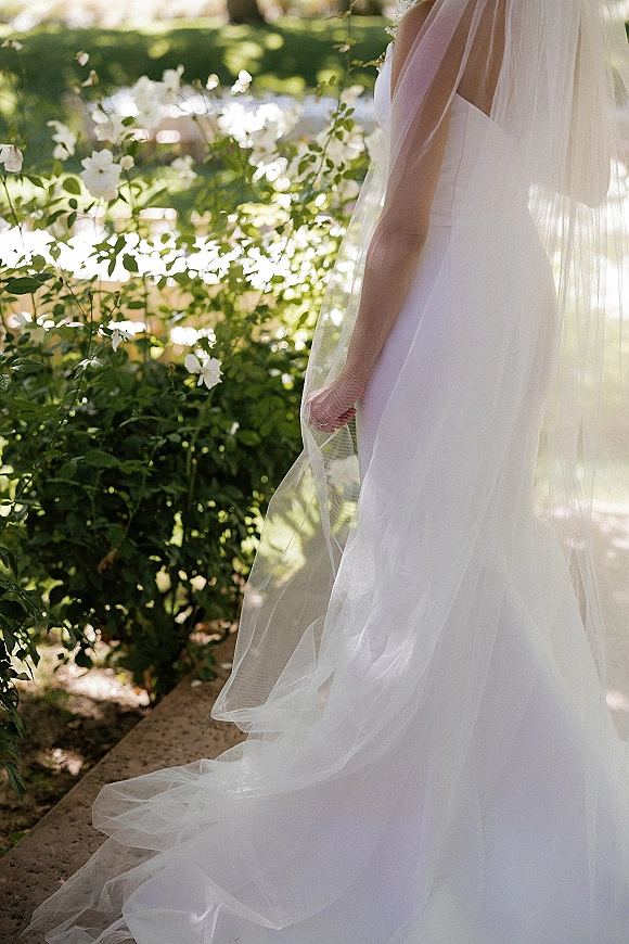 Bridal veil draping in a cathedral length veil over a strapless wedding dress, trailing along a sunlit garden stone path with white flowers
