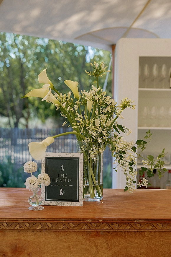 Wedding bar decor with a signature drink menu sign beside a tall glass vase of white calla lilies and greenery on a wood bar under a tent