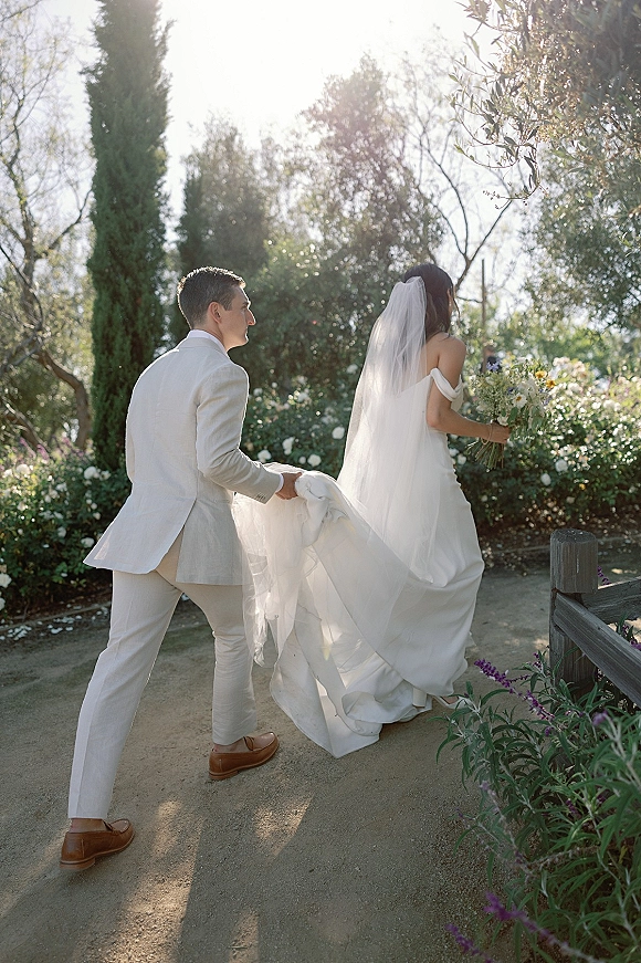 Wedding couple portrait of bride and groom walking away on a sunlit garden path, groom holding her dress train as she carries a bouquet