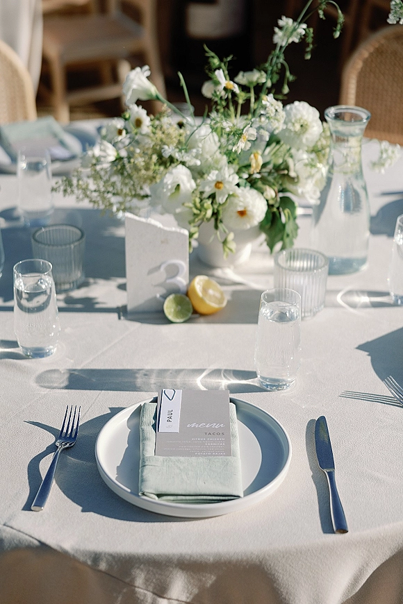 Reception tablescape with white floral centerpiece in ceramic bowl, sage napkin place settings, ribbed votives, and lemon accents in outdoor light