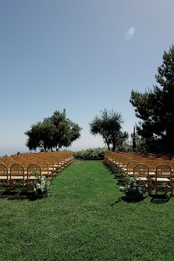 Ceremony setup with wood chairs lined on a grassy aisle, white floral and greenery accents under blue sky and trees