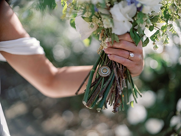 Bridal bouquet of white roses and greenery with long stems, held by bride showing engagement ring and charm locket in sunlit garden