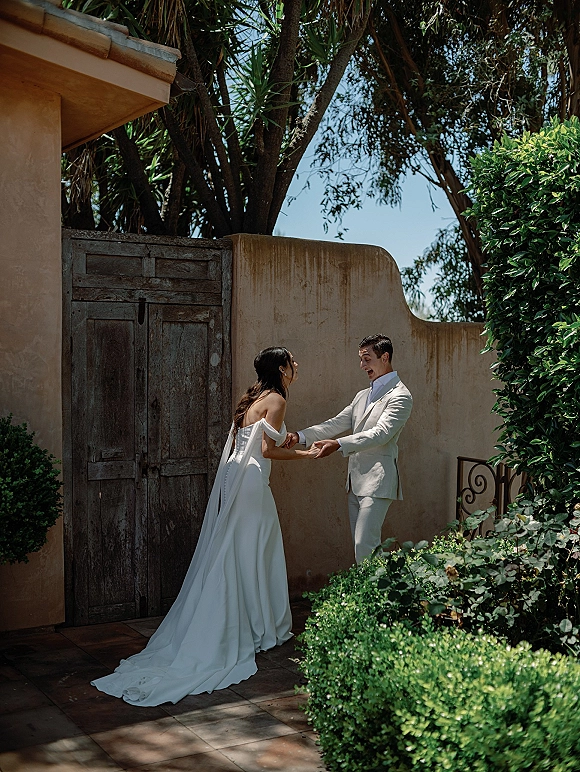 First look moment as the bride approaches the groom, holding hands in an intimate courtyard with a flowing long train behind her