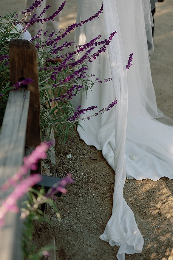 Bridal dress train with long wedding veil trailing on a dirt path beside a wooden fence, accented by small purple flowers