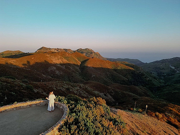 Couple portrait of newlyweds embracing from behind, bride in a wedding dress and groom in suit on a stone terrace at sunset overlook