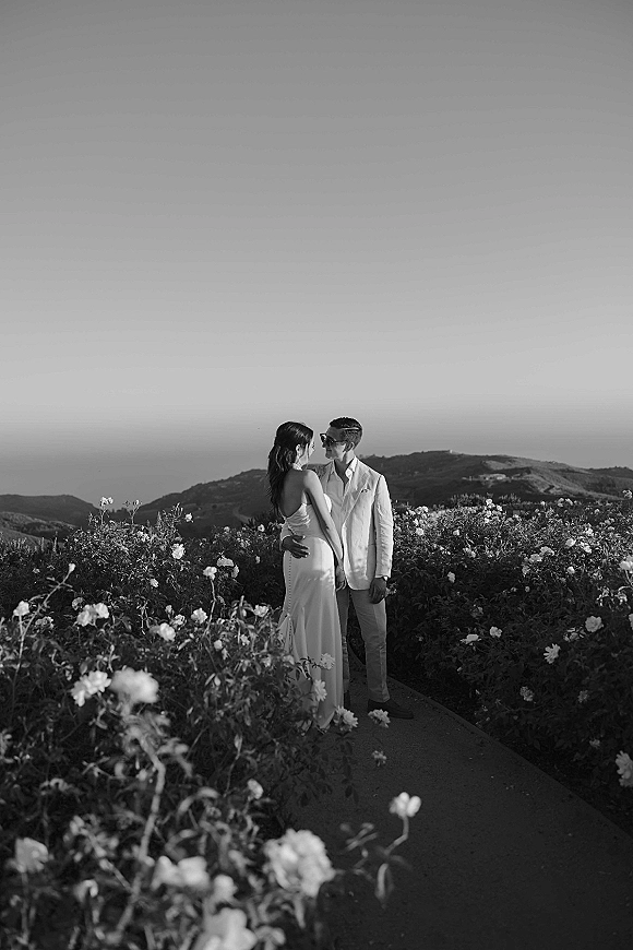 Couple portrait of bride in a backless wedding dress and groom in a light suit with sunglasses, embracing on a path through a flower field with hills and sky