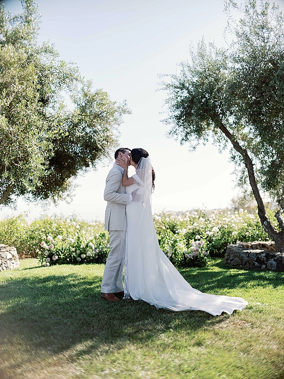 Wedding kiss portrait of bride and groom kissing, her long veil trailing behind, on a garden lawn with olive trees and stone wall backdrop