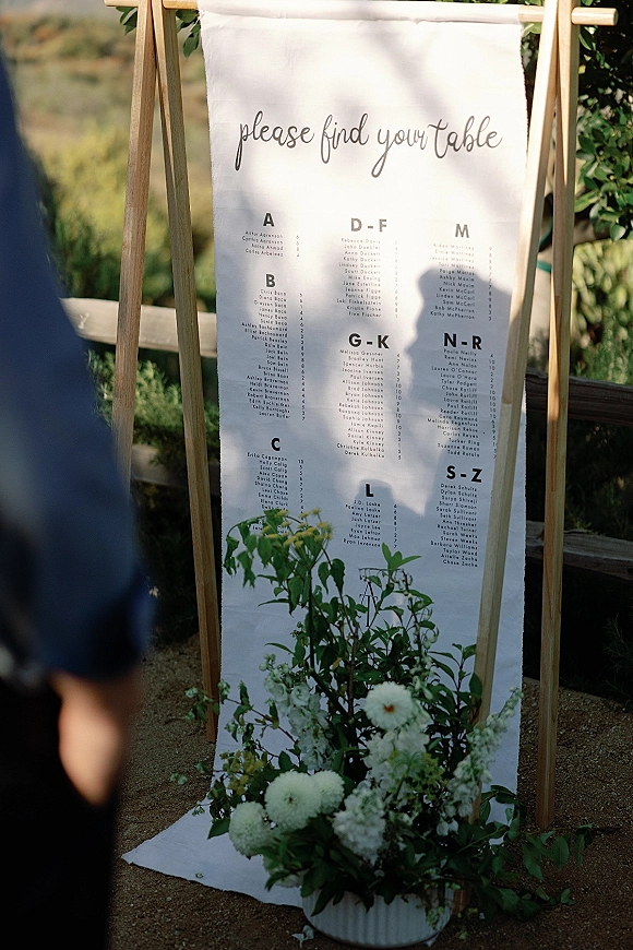Wedding seating chart fabric sign on a wooden easel with calligraphy, a white floral accent, and greenery in an outdoor setting