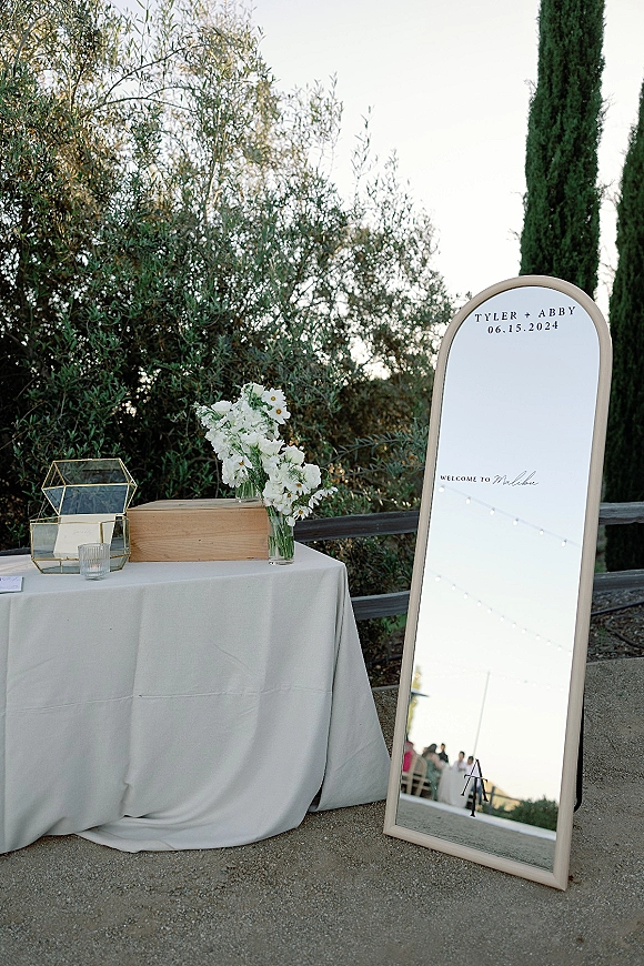 Wedding welcome sign on a full-length mirror with white florals in a glass vase, set on a linen table amid outdoor greenery and fence
