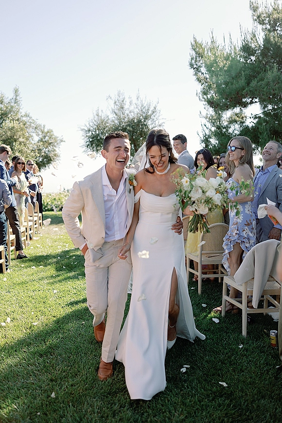 Wedding recessional as bride and groom walk the aisle while guests cheer and toss flower petals on an outdoor lawn, bride holding bouquet and veil flowing