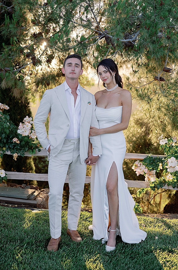 Couple portrait of bride and groom holding hands, bride in strapless slit gown with pearl choker, in sunset garden by pine trees and fence