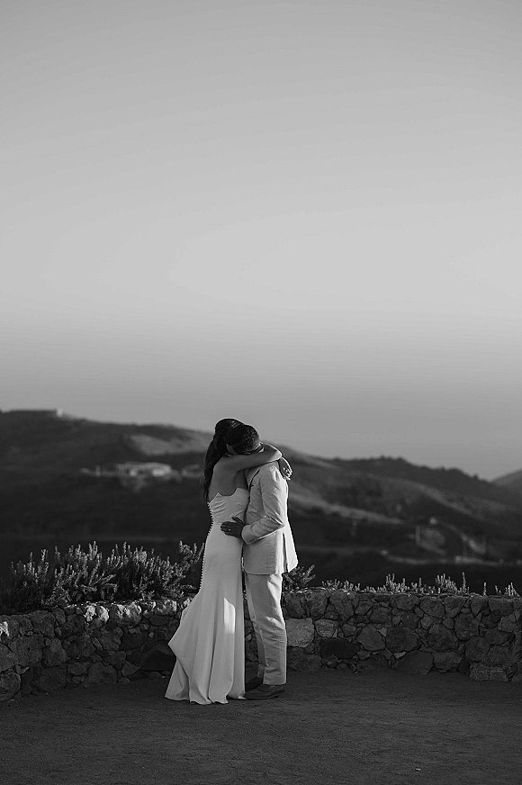 Couple portrait in a black and white wedding portrait, bride in a strapless wedding dress hugging groom by a stone wall with mountain hills behind
