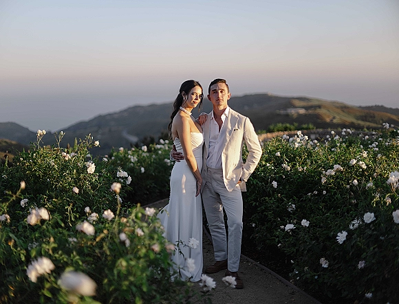 Couple portrait at sunset, bride in strapless dress with pearl choker and groom in white suit on a garden path with ocean and mountains behind