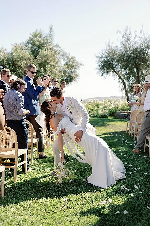 Wedding kiss moment as the bride and groom share a dip kiss in a sunny outdoor aisle, guests in sunglasses tossing rose petals behind