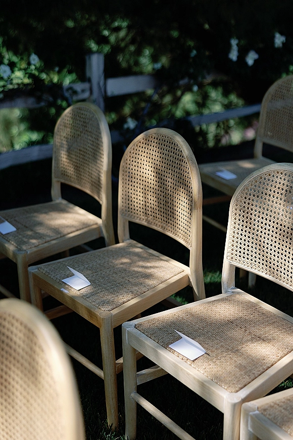 Ceremony seating with outdoor ceremony chairs in neat rows, light wood and cane backs with paper programs beside greenery and fence