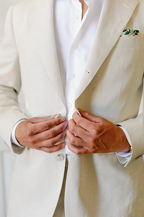 Groom getting ready, buttoning a light tan suit jacket over a white dress shirt, showing cufflinks and pocket square indoors