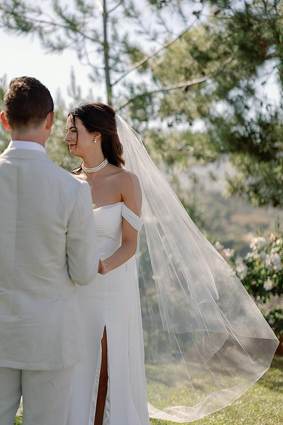 Ceremony moment as bride and groom hold hands during outdoor wedding ceremony, her windy veil and pearl choker on a hillside lawn