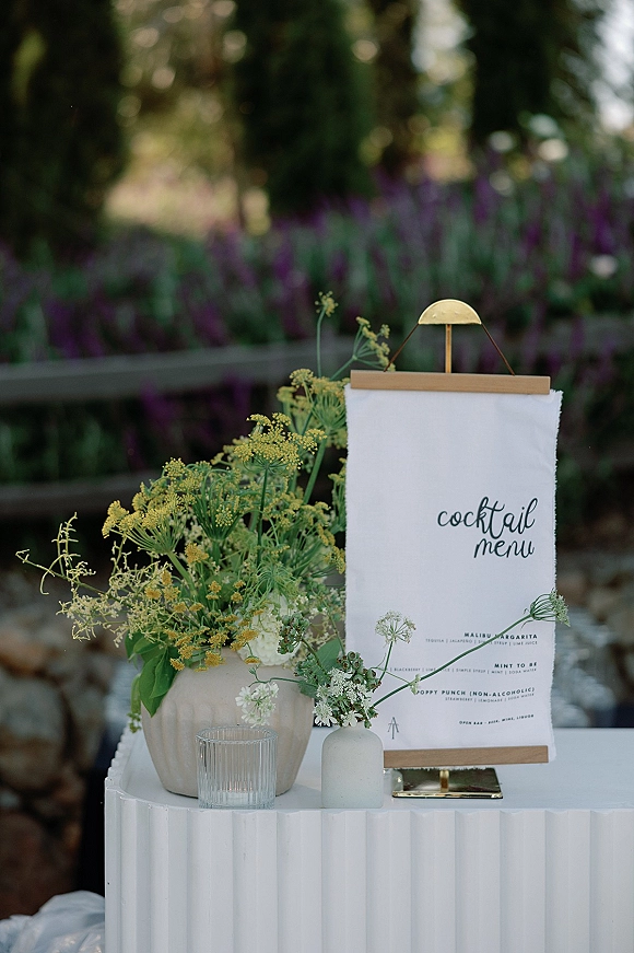 Cocktail menu sign on a tabletop stand with wildflowers and greenery accents beside a votive candle on a garden pedestal table by a wooden fence