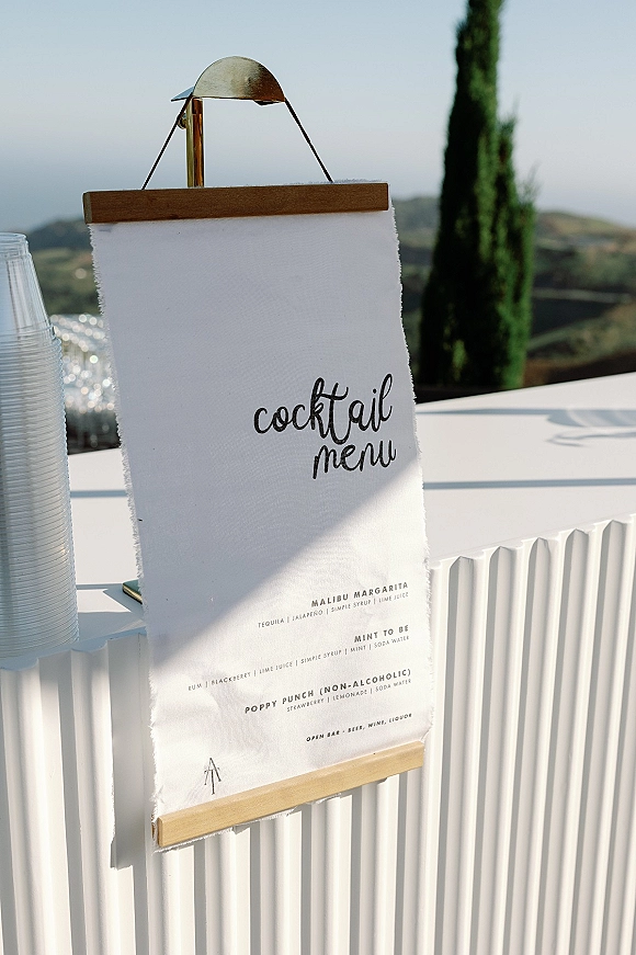 Cocktail menu sign with calligraphy lettering on a linen banner hanging from a wooden hanger at an outdoor bar counter with hills beyond