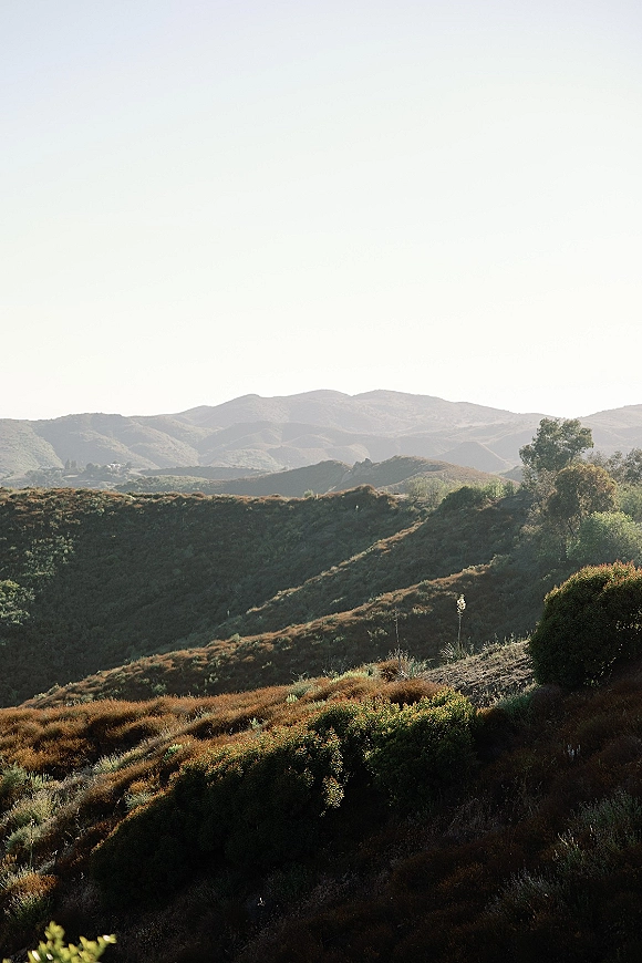 Mountain landscape with rolling hills scenery, shrubs and trees under soft sunlight and a wide open sky fading into hazy ridgelines