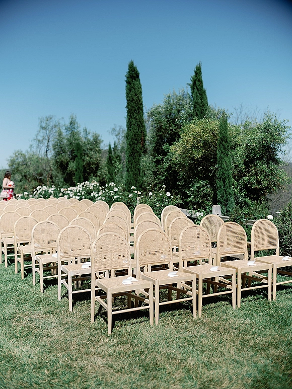 Ceremony seating with cane back wedding chairs and wood chairs in neat rows on a lawn, reserved seat cards facing white roses and cypress trees