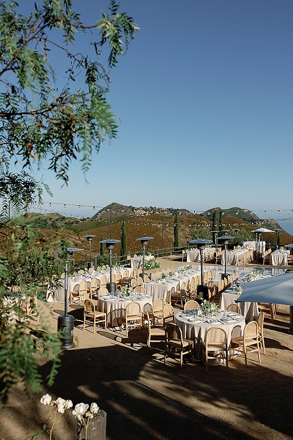 Outdoor reception setup with string lights over white tablecloth long and round tables, rattan chairs, and hillside views under blue sky