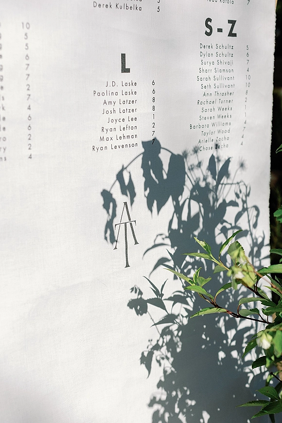 Seating chart sign with printed guest list and monogram, styled in black and white on white paper with soft plant shadows and greenery leaves