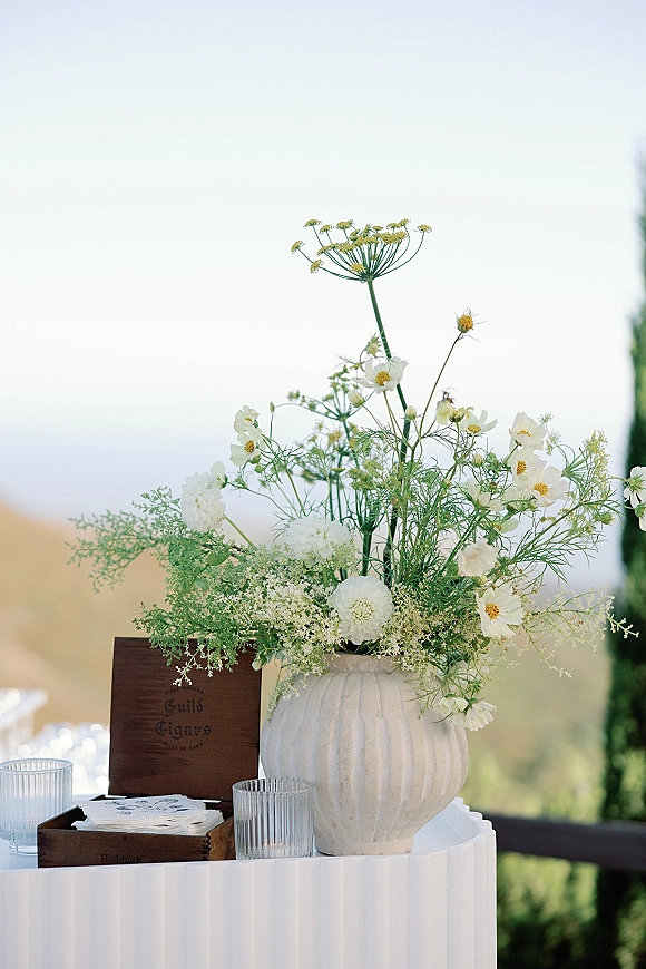 Wedding floral centerpiece with wildflower wedding centerpiece blooms in a white ceramic vase, set beside a wooden cigar box on an outdoor table