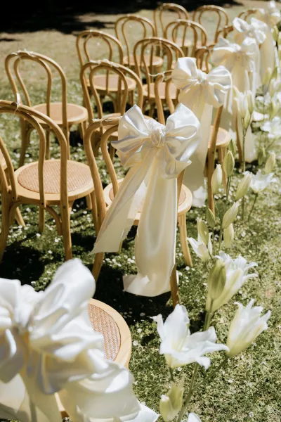 Ceremony aisle decor with wedding aisle flowers of white lilies marking a grassy path beside wood chairs with white satin bows in sunlight