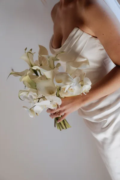Bridal bouquet of white calla lilies and cascading orchids held against a strapless satin wedding dress, ring visible on hand, neutral wall backdrop