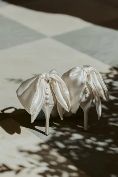 Wedding shoes bridal heels in ivory satin with oversized bows and button detail, resting on stone tiles with sunlit shadows