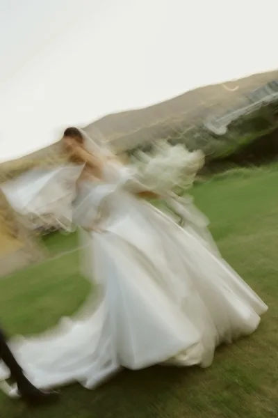 Wedding couple portrait of bride and groom running hand in hand, her veil blowing behind them on a grassy hillside under open sky